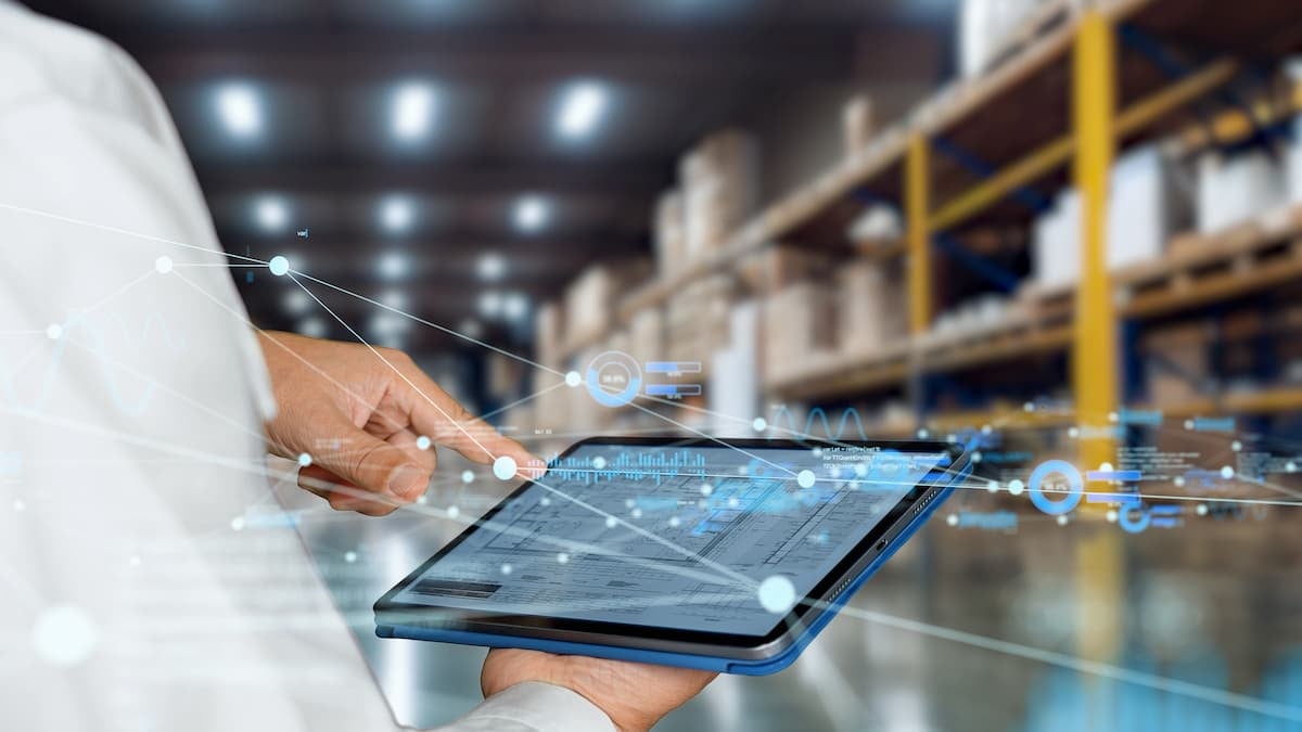 Professional using a tablet in a warehouse environment, with floating digital graphs and data on the screen, symbolizing the use of technology for inventory management and control.