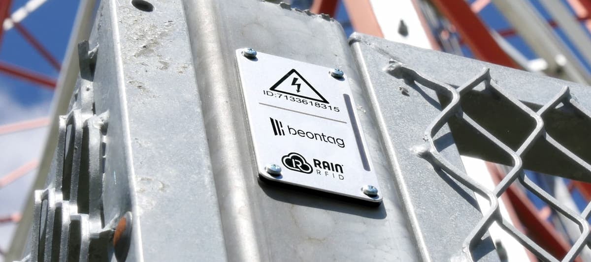 Close-up de uma estrutura metálica com etiqueta RFID (Beontag/RAIN RFID) fixada por parafusos e símbolo de alerta elétrico, com céu azul ao fundo.