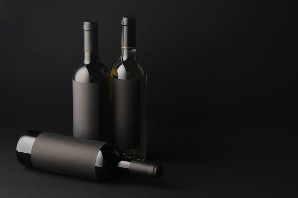 Botellas de vino con etiquetas de papel y BOPP sobre fondo negro, con una botella acostada en primer plano y dos botellas de pie al fondo.