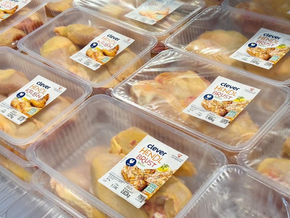 Stacks of plastic trays filled with raw chicken pieces, sealed with clear film and labeled, displayed in a refrigerated store case.