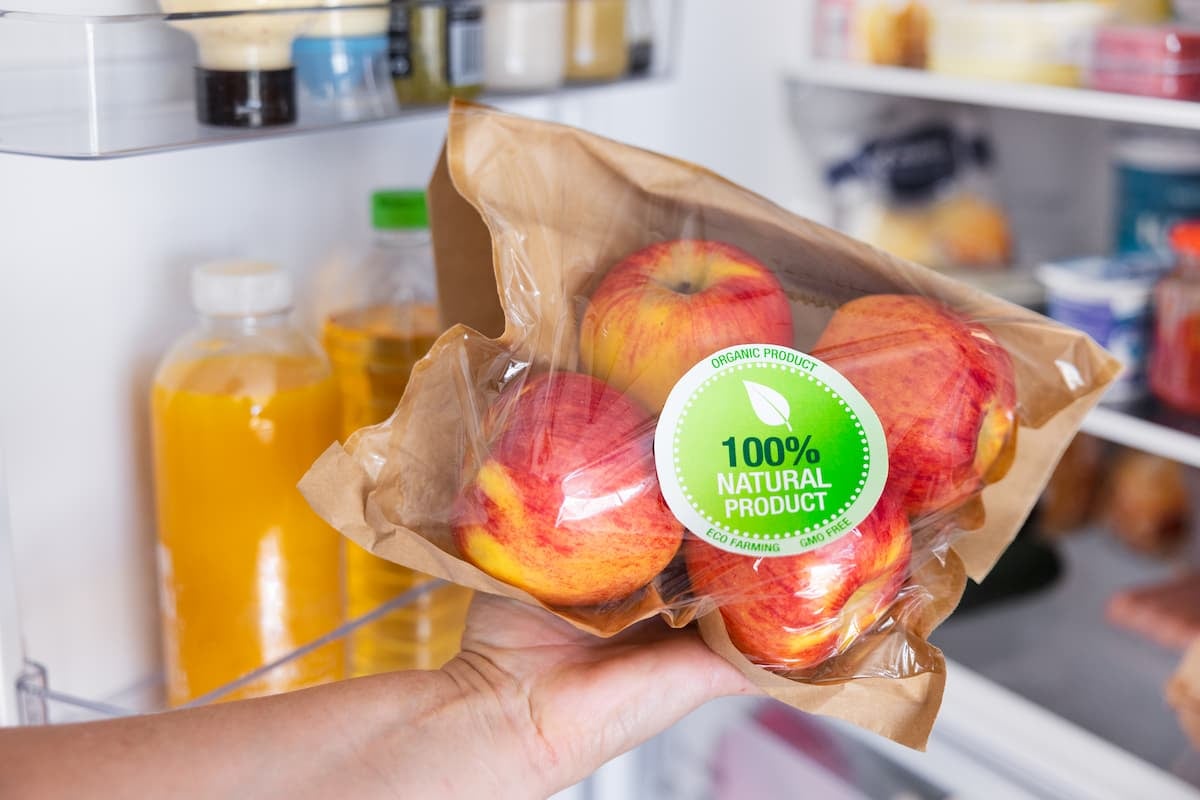 Hand holding a bag of apples with a “100% natural product” sticker inside a refrigerator, highlighting labeling trends in food packaging and consumer transparency.