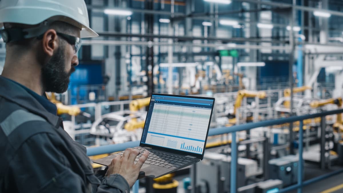 Engineer wearing a safety helmet uses a laptop to monitor production data on an automated factory floor with robotic arms in the background.