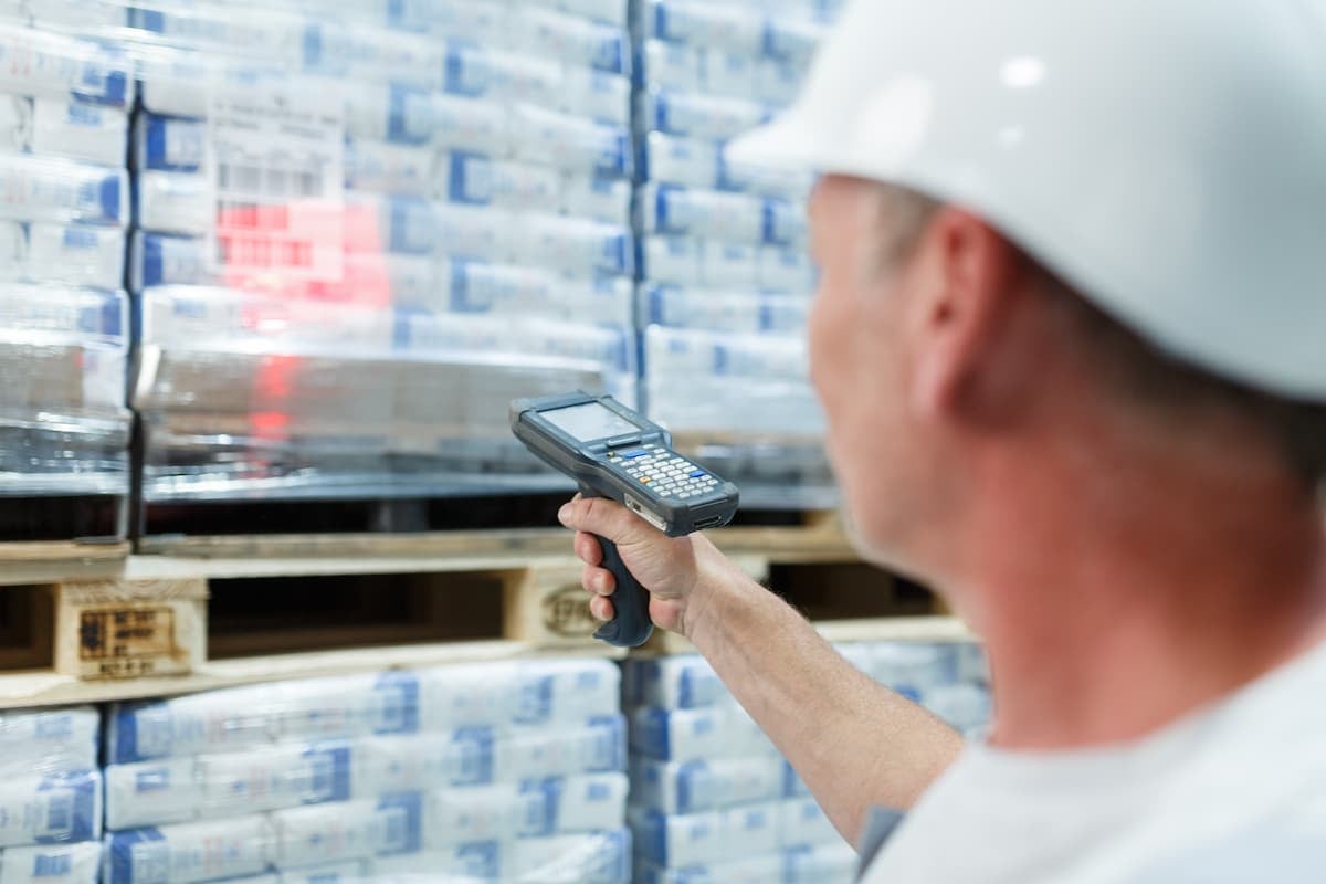 Warehouse worker in a white hard hat scanning a shrink-wrapped pallet with a handheld barcode/RFID reader for inventory tracking.